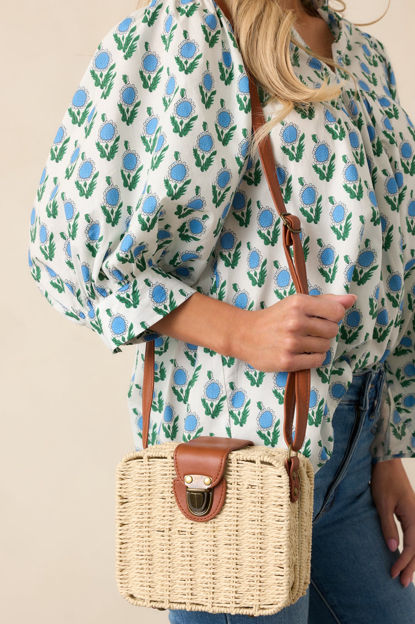 A handbag with a square shape, showcasing the white wicker material, bronze tuck lock closure, and brown faux leather shoulder strap.