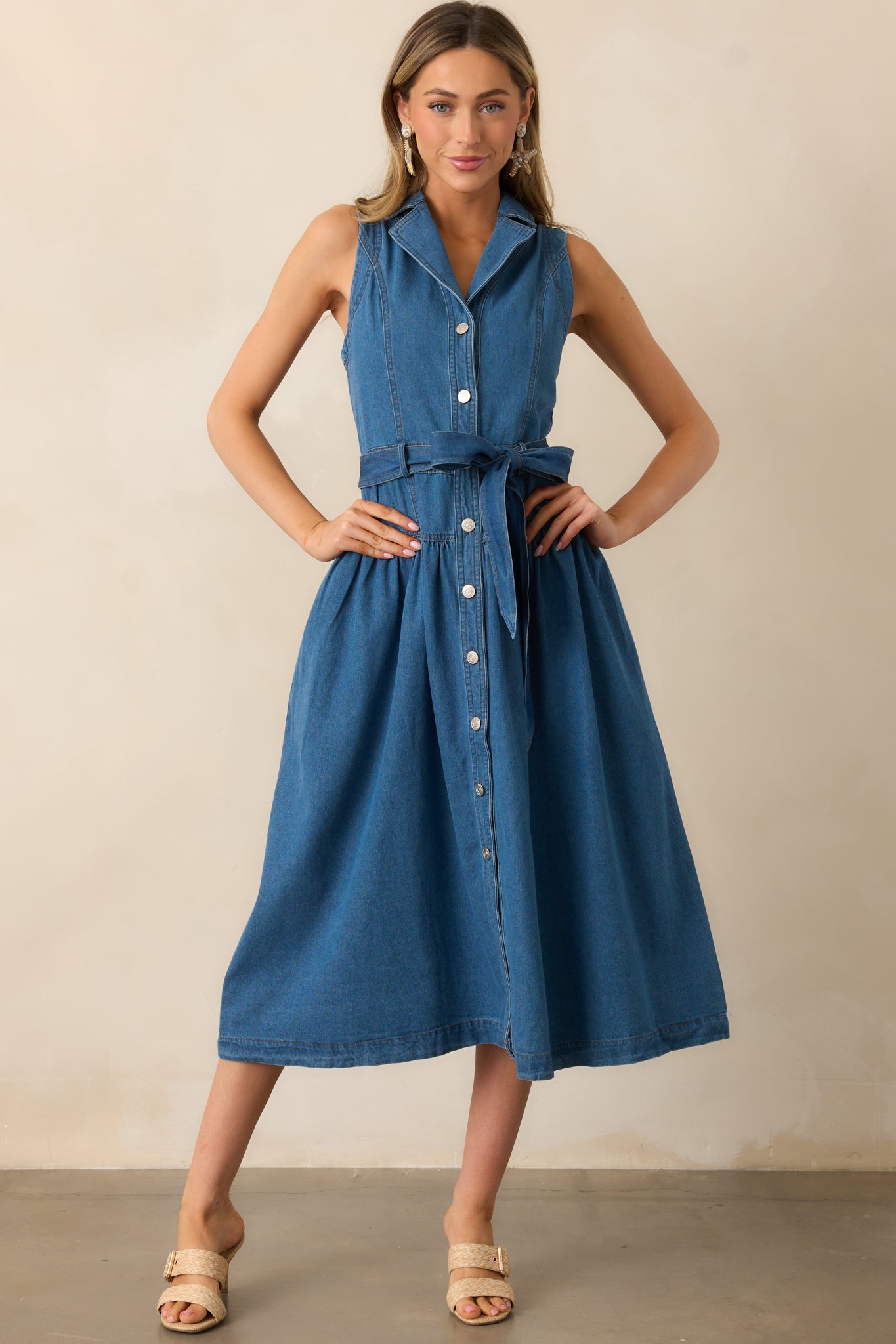 Front-facing shot of the blue denim dress with a midi length, sleeveless cut, and drop waist, accented by a self-tie waistband and button front.