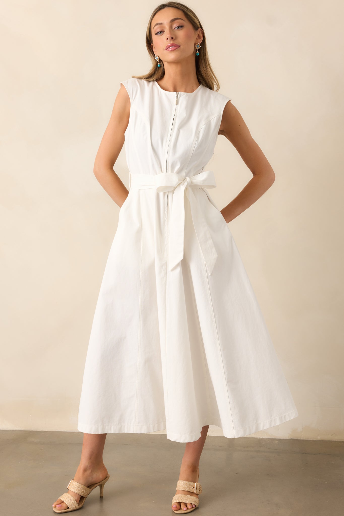 A white midi dress featuring a round neckline, zipper front, visible seams, self-tie waist belt, belt loops, functional hip pockets, and a flowing silhouette.