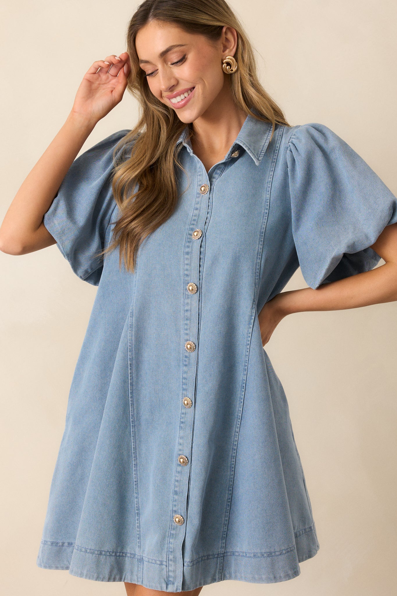 A light wash denim mini dress featuring a collared neckline, functional gold button front, short puff sleeves, and functional pockets for a stylish, casual look.