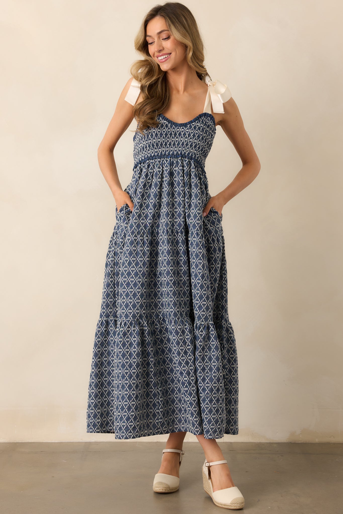 A dark wash midi dress featuring a geometric embroidered pattern, adjustable self-tie bow straps, a fully smocked bust, and a tiered skirt with functional pockets.
