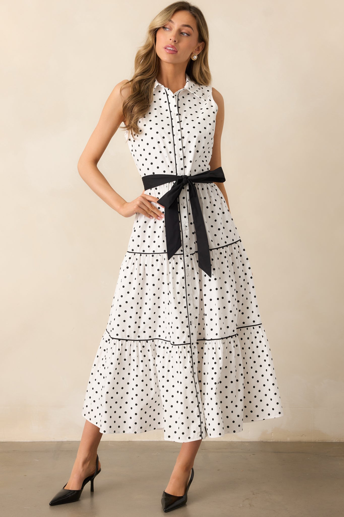 This white dress features a collared neckline, functional button front, and a sleeveless design. Black trim detailing adds contrast throughout, while an optional black self-tie belt enhances the silhouette.
