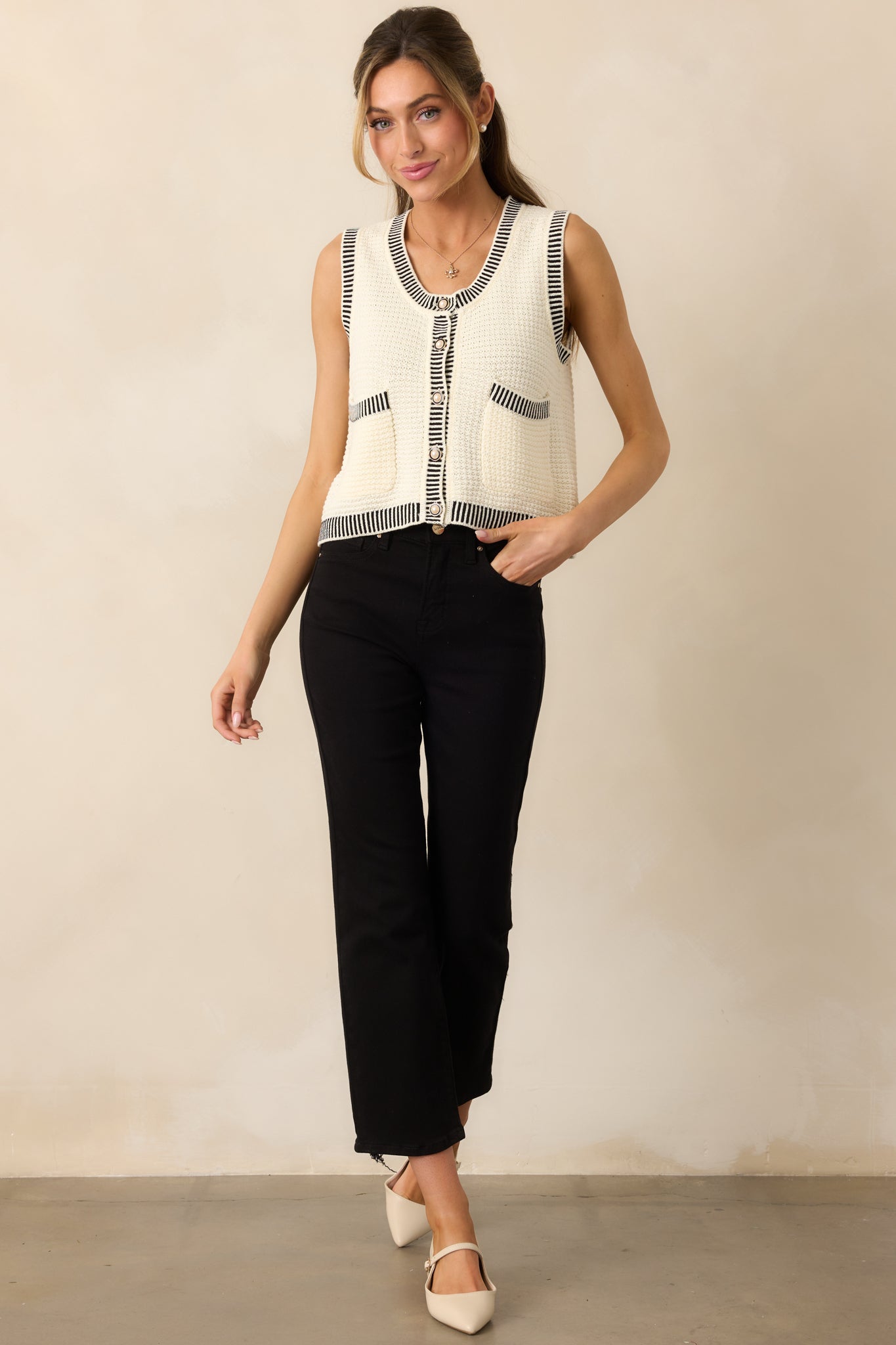 Front view of the ivory sweater vest showcasing the scoop neckline, faux pearl buttons, and black stripe detailing at the hems for a stylish finish.